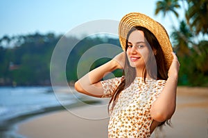 Smiling young woman in straw hat on tropical beach at sunset. Relaxed tourist enjoys warm evening by sea. Leisure in