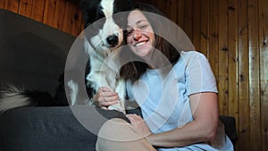 Smiling young woman stroking playing with cute puppy dog border collie on couch sofa at home indoor. Owner girl with pet