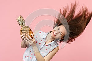 Smiling young woman in summer clothes with fluttering hair hold fresh ripe pineapple fruit isolated on pink pastel wall