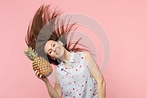 Smiling young woman in summer clothes with fluttering hair hold fresh ripe pineapple fruit isolated on pink pastel wall