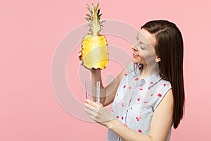 Smiling young woman in summer clothes holding half of fresh ripe pineapple fruit, glass cup isolated on pink pastel wall