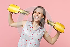 Smiling young woman in summer clothes holding halfs of fresh ripe pineapple fruit isolated on pink pastel wall