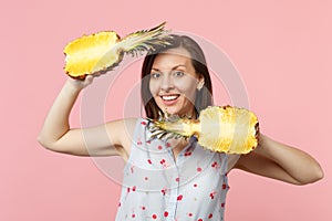 Smiling young woman in summer clothes holding halfs of fresh ripe pineapple fruit isolated on pink pastel wall