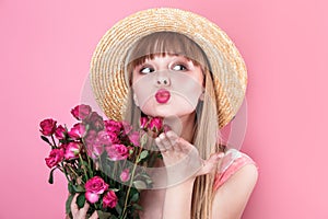 Smiling young woman in summer dress and straw hat holding bouquet of roses and blowing a kiss with hand on air
