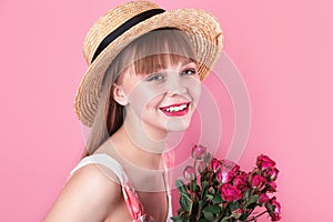 Smiling young woman in summer dress and straw hat holding bouquet of roses on pink background