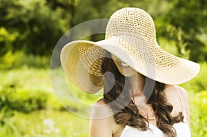 Smiling young woman in sun hat against background of summer green park covering her eyes. Beautiful healthy happy girl