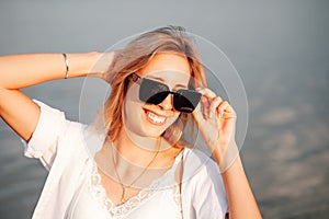 A smiling young woman in sunglasses. Photo of a beautiful young blonde in a white dress and shirt at dawn
