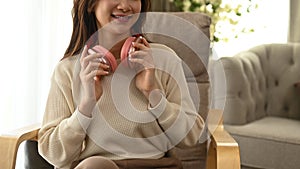 Smiling young woman in sweater drinking resting on armchair, enjoy stress free peaceful mood in cozy living room