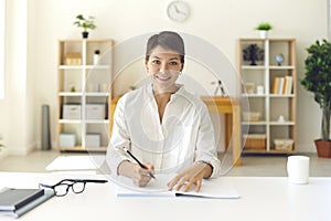 Smiling young woman taking notes while listening to an online lecture in a bright room.