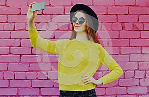 Smiling young woman taking selfie picture by smartphone over a pink brick wall background