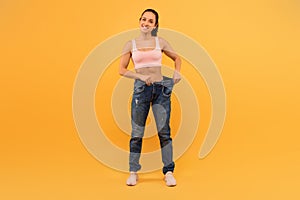 Smiling Young Woman Trying on Oversized Jeans Against Yellow Background