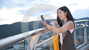 Smiling young woman typing text message on mobile phone while standing on roof terrace outdoors with cityscape on