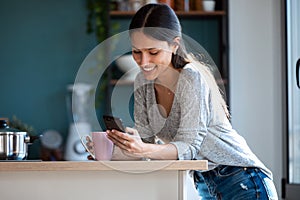 Smiling young woman using her mobile phone while drinking a cup of coffee in the kitchen at home