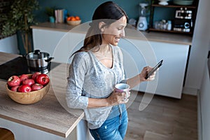 Smiling young woman using her mobile phone while drinking a cup of coffee in the kitchen at home