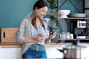 Smiling young woman using her mobile phone while drinking a cup of coffee in the kitchen at home