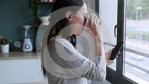 Smiling young woman using her mobile phone while drinking a cup of coffee in the kitchen at home.