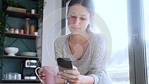 Smiling young woman using her mobile phone while drinking a cup of coffee in the kitchen at home.