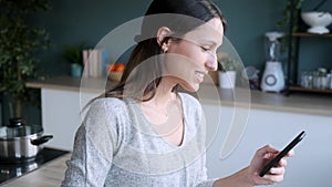 Smiling young woman using her mobile phone while drinking a cup of coffee in the kitchen at home.