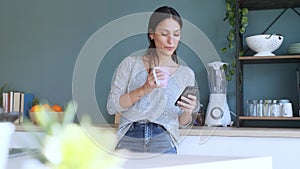 Smiling young woman using her mobile phone while drinking a cup of coffee in the kitchen at home.
