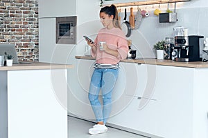 Smiling young woman using her mobile phone while drinking a cup of coffee in the kitchen at home