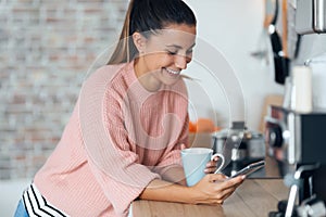 Smiling young woman using her mobile phone while drinking a cup of coffee in the kitchen at home