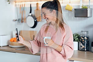 Smiling young woman using her mobile phone while drinking a cup of coffee in the kitchen at home