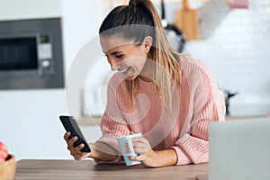 Smiling young woman using her mobile phone while drinking a cup of coffee in the kitchen at home