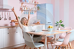 Smiling young woman using laptop in the kitchen at home. Blonde woman works on computer, freelancer or blogger working