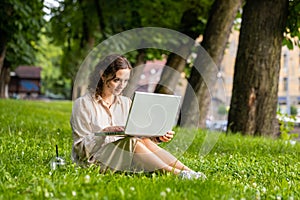 Smiling young woman using laptop typing text answering messages chatting online in city urban park