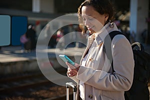 Smiling young woman using mobile phone while waiting train, on the platform of a railway station