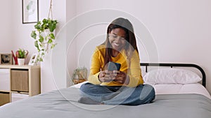 Smiling young woman using smartphone looking at screen while relaxing on bed