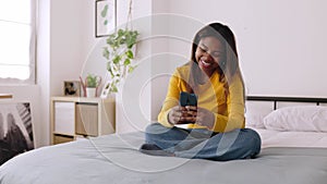 Smiling young woman using smartphone looking at screen while relaxing on bed