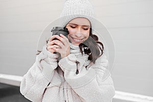 Smiling young woman in warm coat holding cup of coffee outdoors. Woman cofee lover
