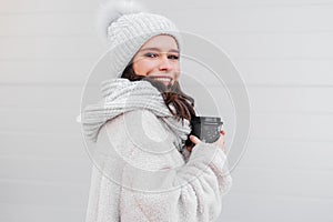 Smiling young woman in warm coat holding cup of coffee outdoors. Woman cofee lover