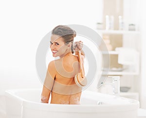 Smiling young woman washing with body brush in bathtub