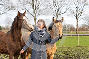 Smiling young woman in a wax coat with her 1 year old stallions in the pasture. She cuddles with them to socialize them