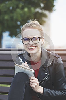 Smiling young woman wearing eyeglasses writing notes on notebook with pen outside, planning day