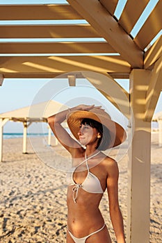 Smiling young woman wearing a hat enjoying her holiday on the beach. Satisfied beautiful girl relaxes on the beach during sunrise