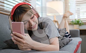Smiling young woman wearing headphones and using smart phone, resting on couch in cozy living room
