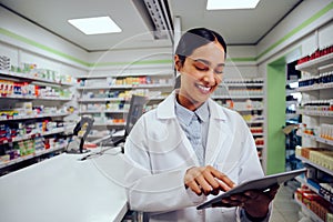 Smiling young woman wearing labcoat standing near counter holding and using digital tablet in chemist