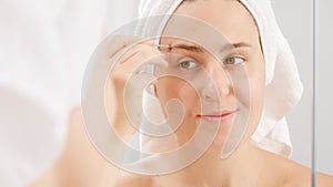Smiling young woman wearing white towel plucking her eyebrows with cosmetic tweezers. Concept of beautiful female, makeup at home
