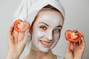 Smiling young woman with white clay mask on face holding fresh tomato against grey background