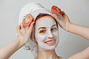 Smiling young woman with white clay mask on face holding fresh tomato against grey background