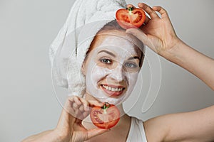 Smiling young woman with white clay mask on face holding fresh tomato against grey background