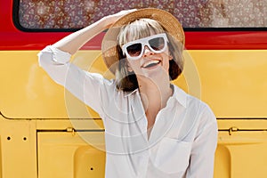 Smiling young woman in in white sunglasses, a straw hat and white shirt poses next to the a bright red and yellow