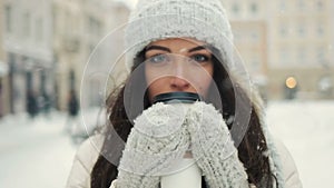 Smiling young woman in white warm clothes with and drinking coffee to take away over snowy city background