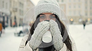 Smiling young woman in white warm clothes with and drinking coffee to take away over snowy city background