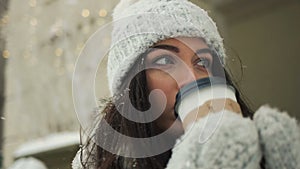 Smiling young woman in white warm clothes with and drinking coffee to take away over snowy city background