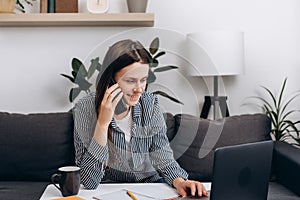 Smiling young woman work on laptop from home office talk on cellphone consulting client or customer online sitting on comfy couch