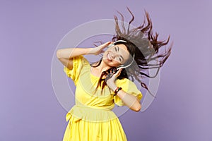 Smiling young woman in yellow dress with flowing hair listen music putting hands on headphones  on pastel violet